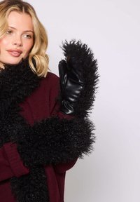 Donna bionda che indossa un maglione bordeaux e una sciarpa di pelliccia finta nera e ricci, mentre tiene un guanto di pelle nera con bordi in pelliccia.
