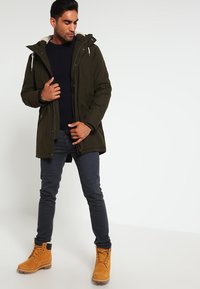 Veste isolante vert olive avec une capuche, dotée d'une doublure en fausse fourrure, d'une fermeture éclair à l'avant et de plusieurs poches, associée à un pull bleu marine et un jean noir.