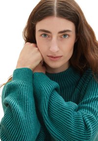 Locker sitzender teal-farbener Pullover mit gerippter Textur und hohem Ausschnitt. Der weiche Stoff umhüllt die Arme und sorgt für ein gemütliches Erscheinungsbild.