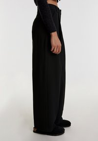 Schwarze weit geschnittene Hose mit glatter Textur, hohem Bund, Fronttaschen und taillierter Passform, kombiniert mit schwarzen Schlüpfschuhen.