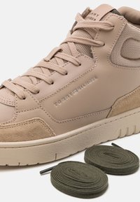 Beige Tommy Hilfiger sneaker met leren en suède panelen, lichtbruine veters en extra opgerolde donkergroene veters naast de schoen.