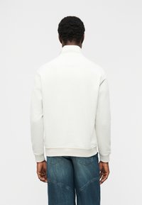 Άνδρας με κοντά σγουρά μαλλιά, φοράει λευκό φούτερ με ψηλό γιακά και μπλε τζιν, γυρισμένος πίσω, με απλό φωτεινό φόντο.