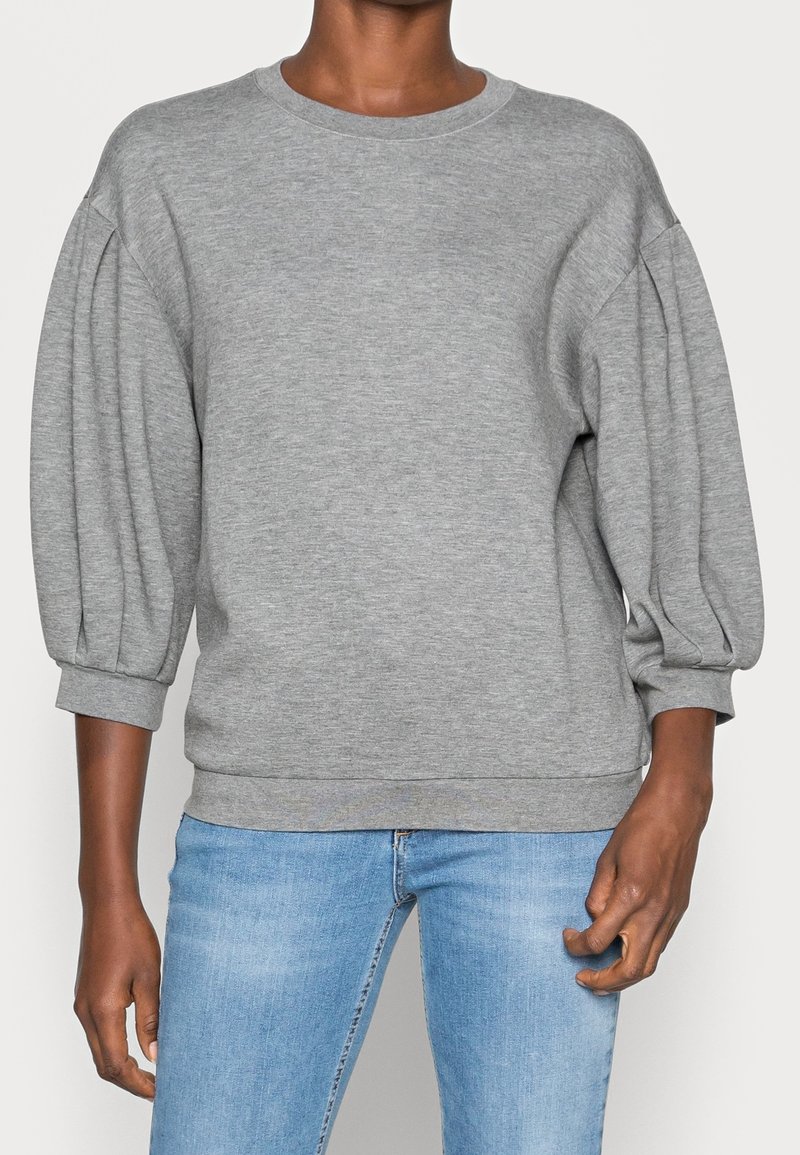 Personne portant un sweat-shirt gris clair avec des manches trois-quarts bouffantes et un jean bleu clair sur un fond uni.