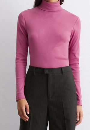 Frau trägt einen eng anliegenden, langärmligen rosa Rollkragenpullover kombiniert mit hoch taillierten schwarzen Nadelstreifenhosen vor einem schlichten Hintergrund.