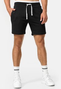 Schwarze Sweat-Shorts aus weichem Material, mit elastischem Taillenbund, weißen Kordeln und Seitentaschen. Knielange Ausführung.