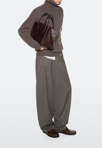 Persona con maglione marrone a collo alto, pantaloni larghi grigi, scarpe marroni, che tiene sotto il braccio una borsa in pelle testurizzata marrone scuro.