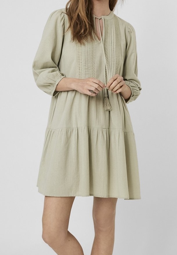 Femme portant une robe beige clair, ample, longueur genou, manches longues, taille froncée et lien à pompons au niveau de l'encolure.