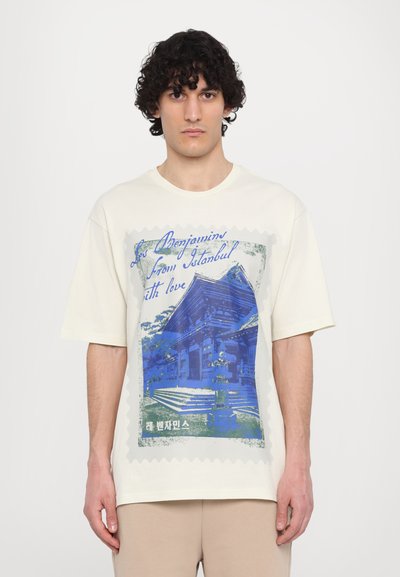 Κρέμμα χρώματος oversized μπλουζάκι με μπλε γραφικό σχέδιο ενός κτηρίου, περιβεβλημένο με σχέδιο γραμματόσημου και κείμενο σε μπλε και πράσινο.