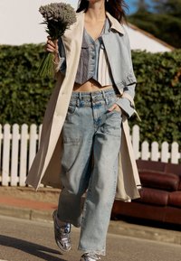 Jasnokremowy prochowiec nałożony na skróconą niebieską kurtkę dżinsową, w połączeniu z wysokim stanem niebieskich dżinsów i srebrnymi sneakersami, trzymając bukiet kwiatów.