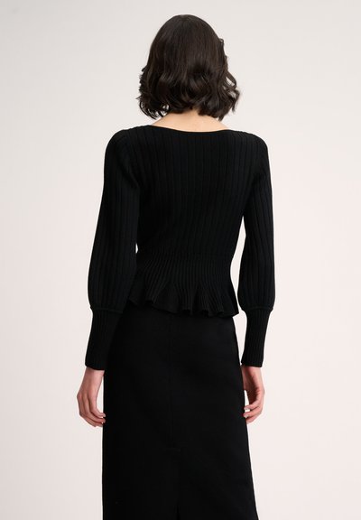 Schwarzer gerippter Strickpullover mit langen Ärmeln, einer Peplum-Taille und einem runden Rückenausschnitt. Getragen mit einem passenden schwarzen Rock mit einem vorderen Schlitz.