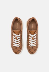 Paire de baskets marron avec des lacets blancs, ornées de motifs de cercles noirs et de croix, vues de dessus sur un fond blanc.