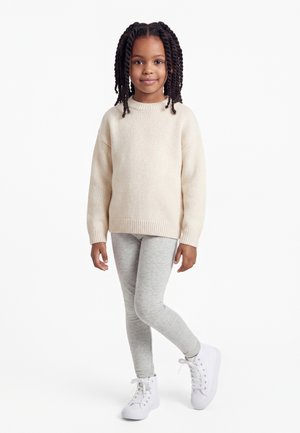 Mlada deklica s pletenicami, oblečena v kremasto pulover, svetlo sive pajkice in bele superge z visokim zgornjim delom, stoji z eno nogo prekrižano.