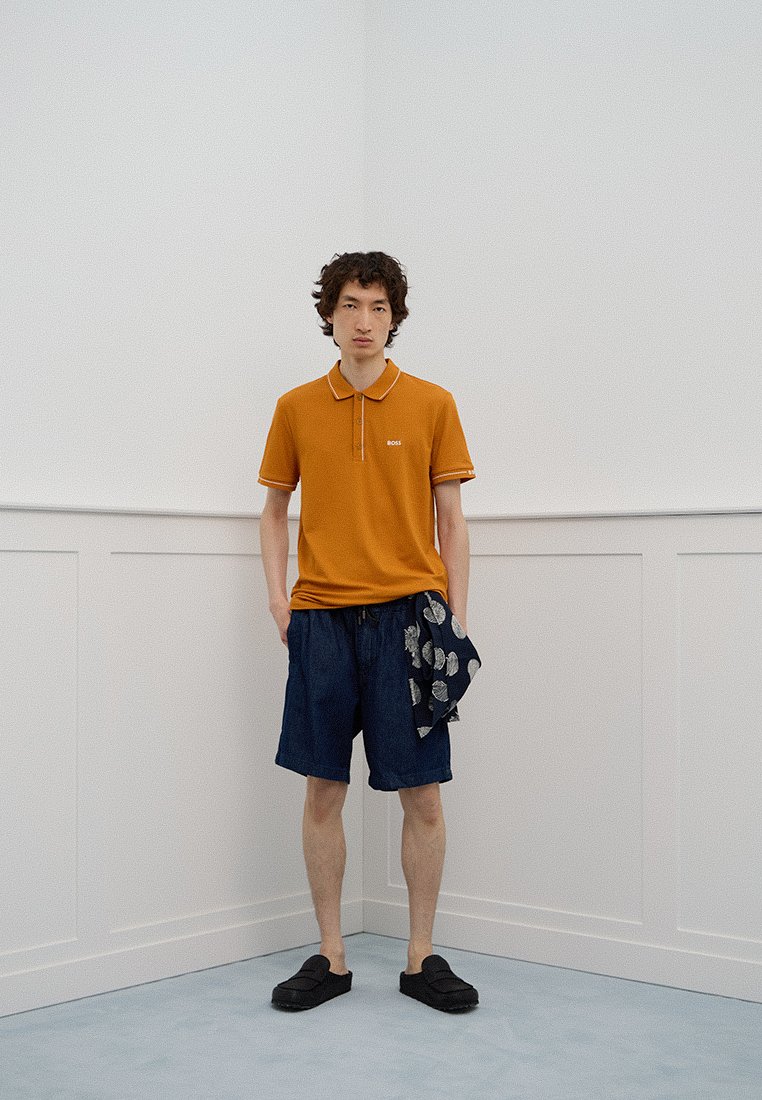 Un giovane uomo sta in un angolo indossando una polo arancione, pantaloni corti blu scuro, scarpe nere, tenendo una sciarpa blu scuro con motivi bianchi in una mano.