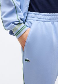 Άτομο που φοράει ανοιχτό μπλε αθλητικό παντελόνι και μπουφάν με πράσινα περιθώρια, χέρι στην τσέπη του παντελονιού, ορατό το λογότυπο κροκόδειλου στο παντελόνι.