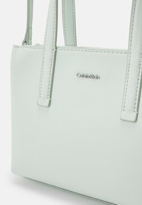 Borsa a tracolla sintetica verde menta con doppi manici, caratterizzata da un accento logo in argento e dettagli cuciti lungo i bordi. Forma rettangolare compatta.