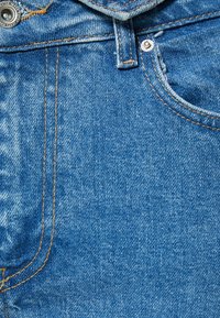 Tela de mezclilla azul con un acabado texturizado, costuras naranjas en contraste, un botón de metal y un borde de bolsillo limpio.