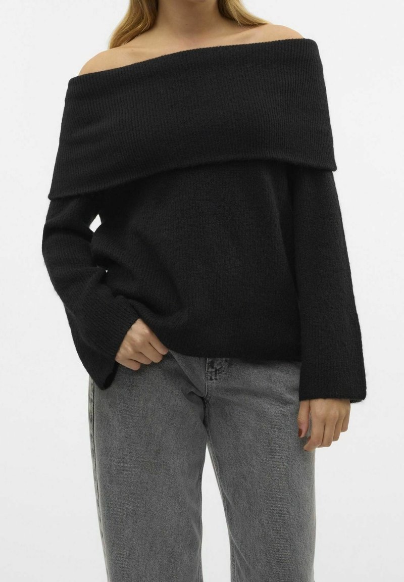 Pull noir à épaules dénudées en texture côtelée avec un large col et des manches longues, associé à un jean gris clair.