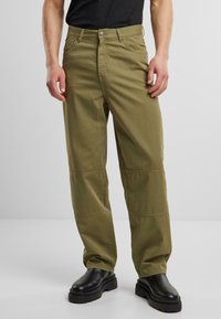 Pantalones cargo de algodón caqui con bolsillos laterales, corte recto, costuras reforzadas en las rodillas y cierre de botón. Combinados con botas negras de suela gruesa.
