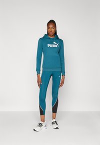 Felpa teal con un grande logo Puma bianco, abbinata a leggings teal con accenti neri e piping bianco. Indossata con sneakers bianche.