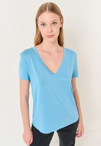 Jasnoniebieska koszulka z dekoltem w serek i krótkimi rękawami, wykonana z miękkiej tkaniny, o luźnym kroju i zaokrąglonym dołem.