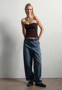 Top a bretelle marrone a coste e jeans denim blu larghi con una cintura marrone, accessoriato con orecchini rotondi, su uno sfondo neutro.