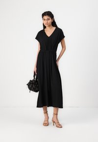 Black maxi dress with short sleeves and a V-neck, featuring a tied waist. Paired with a textured handbag and strappy high-heeled sandals.