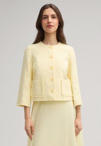 Giacca gialla con texture e scollo rotondo, bottoni frontali e due tasche anteriori. Presenta maniche a tre quarti con bordi sfilacciati.