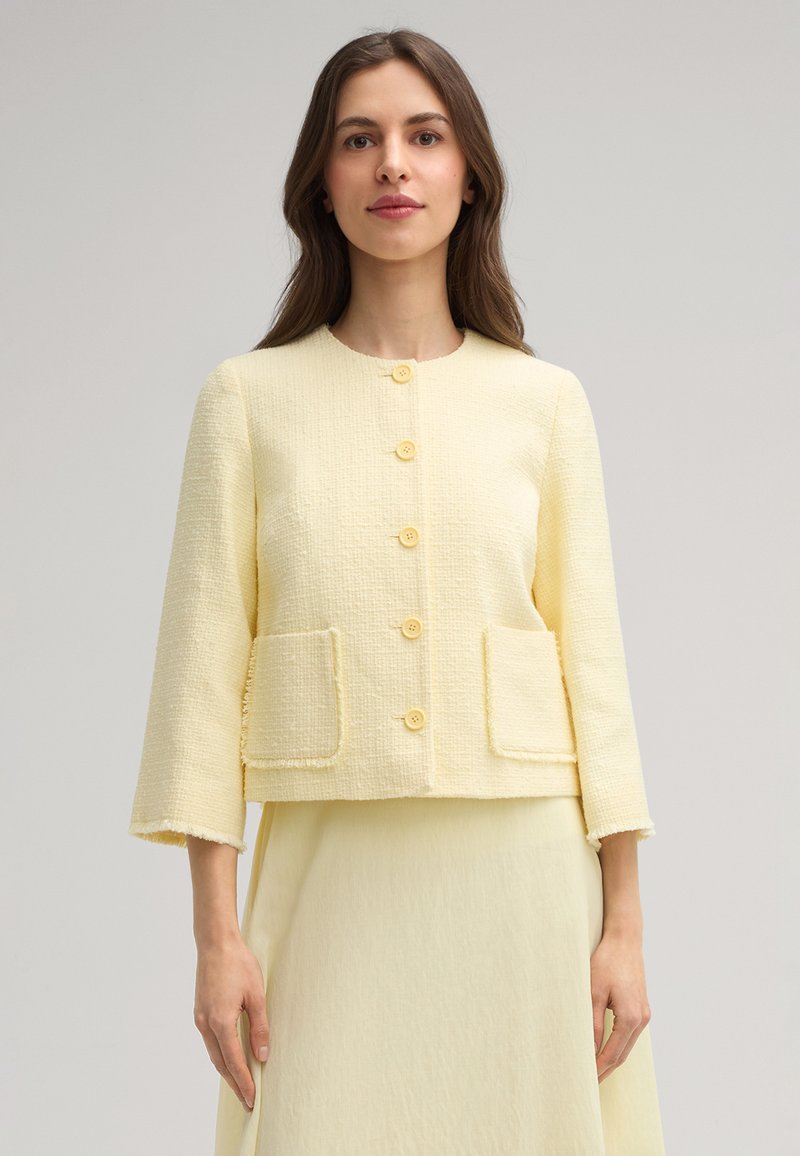 Giacca gialla con texture e scollo rotondo, bottoni frontali e due tasche anteriori. Presenta maniche a tre quarti con bordi sfilacciati.