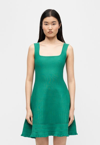 Femme aux longs cheveux bruns portant une robe sans manches verte côtelée avec un décolleté carré et un ourlet à volants, debout devant un fond uni.