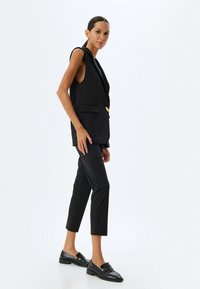 Schwarze ärmellose Blazer mit Reverskragen und goldenem Knopf, kombiniert mit schwarzen, taillierten Hosen und schwarzen Slippern, vor einem weißen Hintergrund.