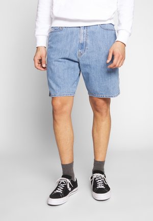 Homme portant un short en denim bleu clair, un sweat-shirt blanc, des chaussettes grises et des baskets noires, debout contre un fond uni.