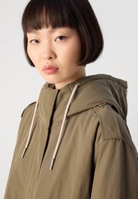 Ελαφρύ μπουφάν ελιάς με κουκούλα, που διαθέτει πλήρες φερμουάρ, κορδόνια και επιθέματα στους ώμους για επιπλέον λεπτομέρεια.