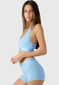 Šviesiai mėlynas liemenėlės ir šortų rinkinys, pagamintas iš minkštos medžiagos, pasižymintis besiūliu dizainu ir logotipo juosta. Lygi tekstūra, prigludimas ir kasualus stilius.