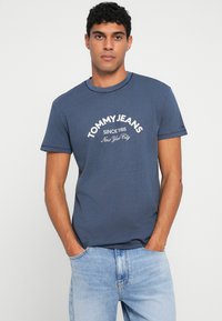 Jaunas vyras, apsirengęs tamsiai mėlyna "Tommy Jeans" marškinėliais ir šviesiai mėlynomis džinsomis, stovi su rankomis kišenėse prieš paprastą foną.