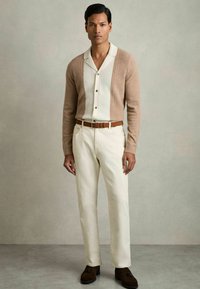 Cardigan en tricot beige sur chemise blanche à boutons, pantalon crème et ceinture en cuir marron. Chaussures marron foncé avec des accents.