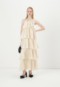 Long, cream-colored dress made of layered, glossy fabric with a halter neckline. Model wears black heeled shoes and a small black handbag.