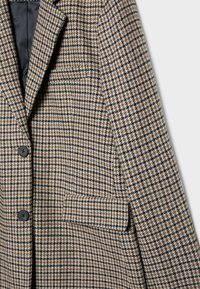 Blazer à motif pied-de-poule marron et beige avec un col cranté, deux boutons noirs, et une poche poitrine. Doublure intérieure lisse visible.