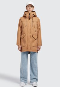 Tanmatterne wasserdichte Parka-Jacke mit verstellbarem Kapuzenzug, zwei Fronttaschen und Reißverschluss, kombiniert mit hellblauen, weit geschnittenen Jeans und weißen Sneakers.