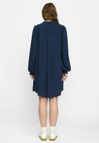Dunkelblaues Langarmkleid mit plissiertem Rücken, aus weichem Stoff und mit elastischen Bündchen. Getragen mit weißen Socken und hellen Schuhen.
