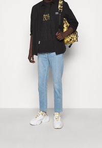 Schwarzes Hemd über schwarzem Grafik-T-Shirt, hellblaue gerade geschnittene Jeans und weiße Sneaker mit goldenen Akzenten. Gelber Rucksack mit Muster.