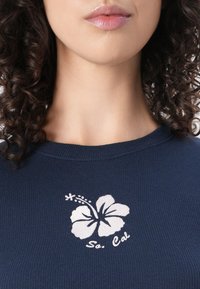 Σκούρο μπλε ριγέ μπλουζάκι με λευκή κεντημένη ίμπισκους λουλούδι και κείμενο "So. Cal". Στρογγυλή λαιμόκοψη και εφαρμοστή σχεδίαση.