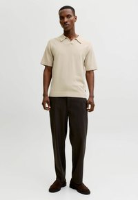 Polo en maille beige avec col, manches courtes, associé à un pantalon plissé marron foncé et des chaussures en suède marron. Design et coupe simples.