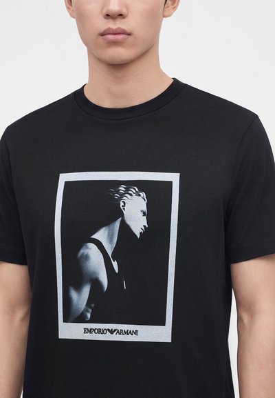 Νέος άνδρας που φοράει μαύρο T-shirt με ασπρόμαυρο γραφικό προφίλ προτομής ενός ατόμου μέσα σε λευκό ορθογώνιο πλαίσιο.