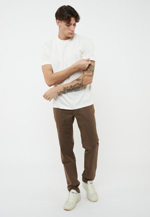 Giovane uomo che sistema la manica del braccio destro tatuato, indossa una maglietta bianca, pantaloni marroni e sneaker bianche, in piedi davanti a uno sfondo semplice.