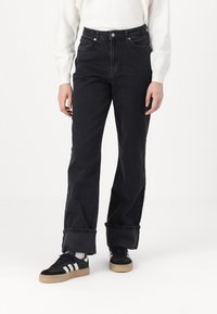 Black denim straight-leg jeans feature a high waist, visible stitching, and rolled cuffs. Paired with black and white athletic shoes.