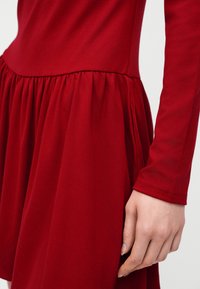 Το χέρι μιας γυναίκας ακουμπά δίπλα σε ένα κόκκινο φόρεμα με μακριά μανίκια, εφαρμοστό μπούστο και φουσκωτή φούστα.