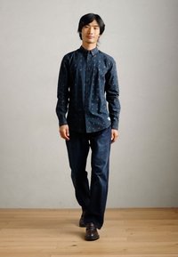 Σκούρο μπλε πουκάμισο με μικρά πολύχρωμα σχέδια, συνδυασμένο με σκούρες τζιν παντελόνες και καφέ παπούτσια, στέκεται σε ξύλινο πάτωμα.