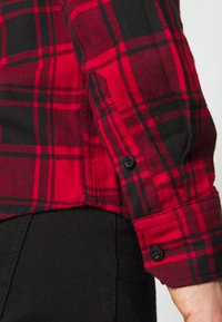 Rot-schwarz karierte Flanellhemd mit strukturierter Oberfläche, ausgestattet mit schwarzen Knöpfen an den Manschetten und einem geraden Saum.