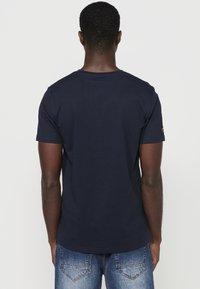 Camiseta de algodón azul marino con mangas cortas, escote redondo, textura suave; presenta un pequeño emblema en el hombro izquierdo.