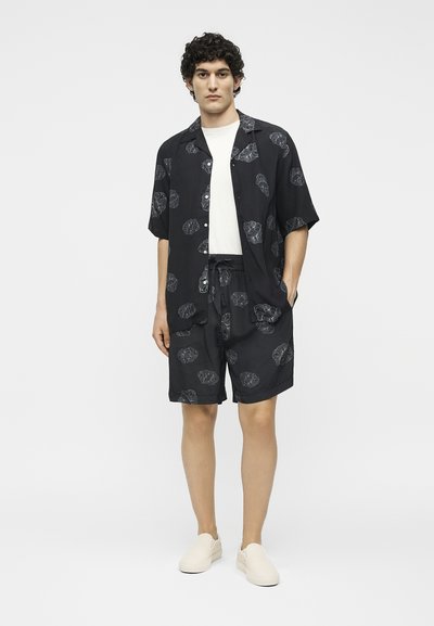 Νέος άνδρας με μαύρο κοντομάνικο πουκάμισο με λουλούδια και ταιριαστό σορτς πάνω από λευκή μπλούζα, λευκά παπούτσια slip-on, στέκεται με το ένα χέρι στην τσέπη.